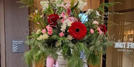 A tall, elegant floral arrangement featuring red gerbera daisies, pink lilies, and greenery in a silver vase.