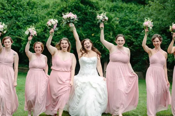 A bride and bridesmaids in pink dresses holding up floral bouquets outdoors.