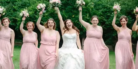 A bride and bridesmaids in pink dresses holding up floral bouquets outdoors.
