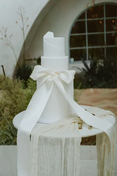 A white three-tier wedding cake with a large ribbon bow displayed on a pedestal table outdoors.