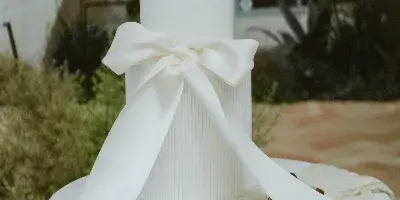 A white three-tier wedding cake with a large ribbon bow displayed on a pedestal table outdoors.