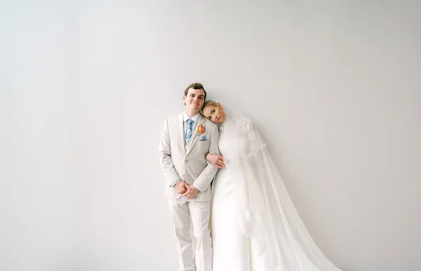 A bride and groom posing together in a white studio setting, with the bride wearing a veil and the groom in a light-colored suit.