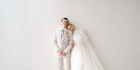 A bride and groom posing together in a white studio setting, with the bride wearing a veil and the groom in a light-colored suit.