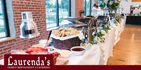 A catering buffet table featuring chafing dishes, a basket of rolls, and a brick wall background, with the Laurenda's Family Restaurant & Catering logo in the corner.