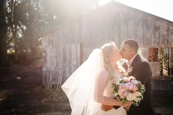 A bride and groom sharing a kiss outdoors in front of a rustic wooden barn, with the bride holding a bouquet of pink and white roses.