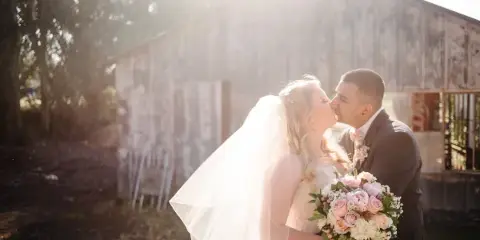 A bride and groom sharing a kiss outdoors in front of a rustic wooden barn, with the bride holding a bouquet of pink and white roses.
