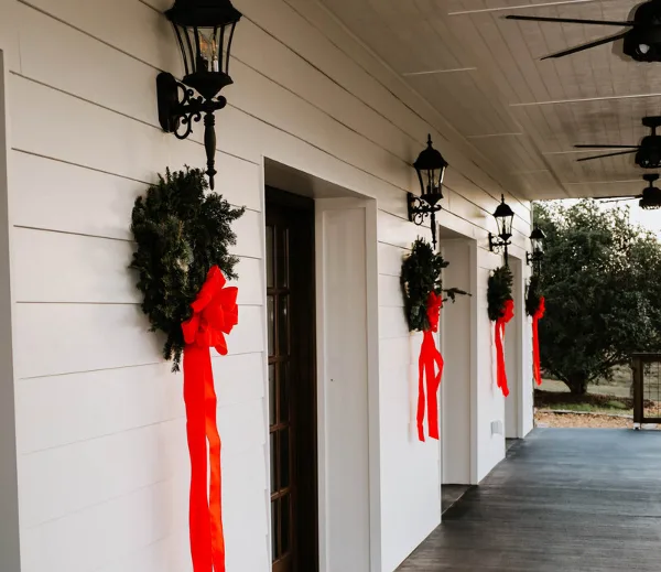 White venue porch with black lanterns, holiday wreaths with red ribbons, and outdoor greenery visible in the background.