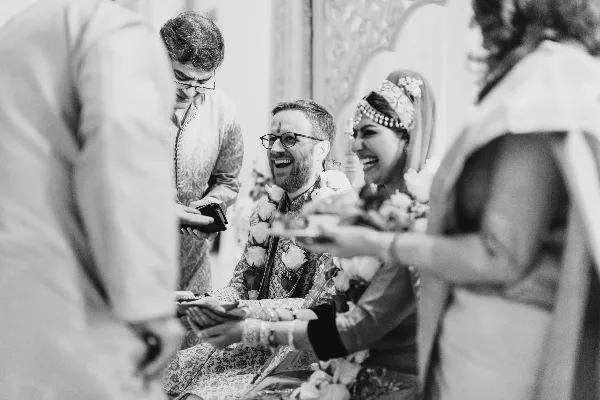 A black and white photograph of a smiling couple during a wedding ceremony, with the bride wearing traditional headwear and the groom wearing a floral garland.