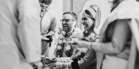A black and white photograph of a smiling couple during a wedding ceremony, with the bride wearing traditional headwear and the groom wearing a floral garland.