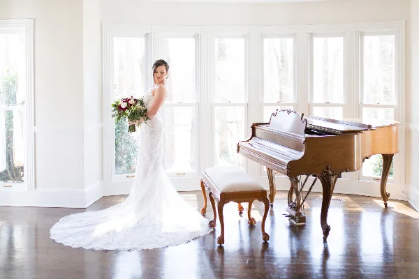 A bride in a white wedding dress stands holding a bouquet next to a grand piano in a bright room with large windows.