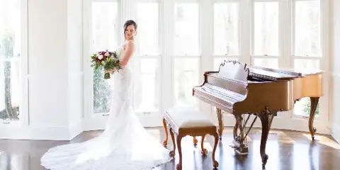 A bride in a white wedding dress stands holding a bouquet next to a grand piano in a bright room with large windows.