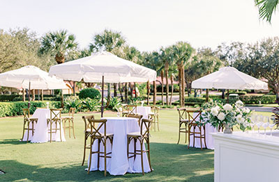 Outdoor event setup with white umbrellas, tables, and floral arrangements on a lawn.