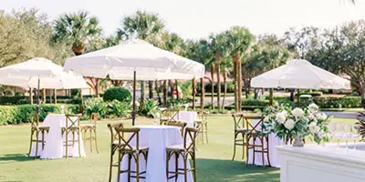 Outdoor event setup with white umbrellas, tables, and floral arrangements on a lawn.
