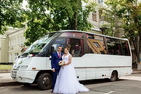 A bride and groom posing in front of a white minibus decorated with flowers.