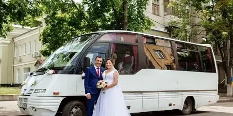 A bride and groom posing in front of a white minibus decorated with flowers.
