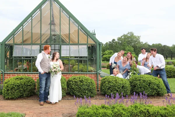 A bride and groom stand with their wedding party in front of a glass greenhouse structure at a garden venue.