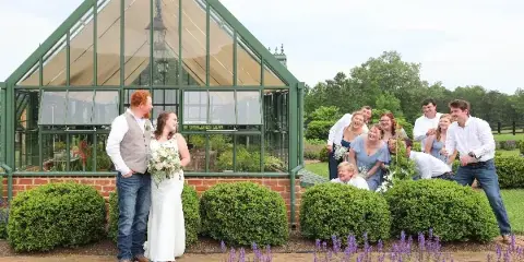 A bride and groom stand with their wedding party in front of a glass greenhouse structure at a garden venue.