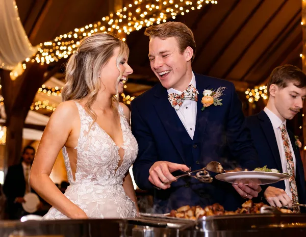 A bride and groom smiling while serving food from a buffet at a wedding reception.