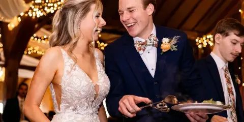 A bride and groom smiling while serving food from a buffet at a wedding reception.