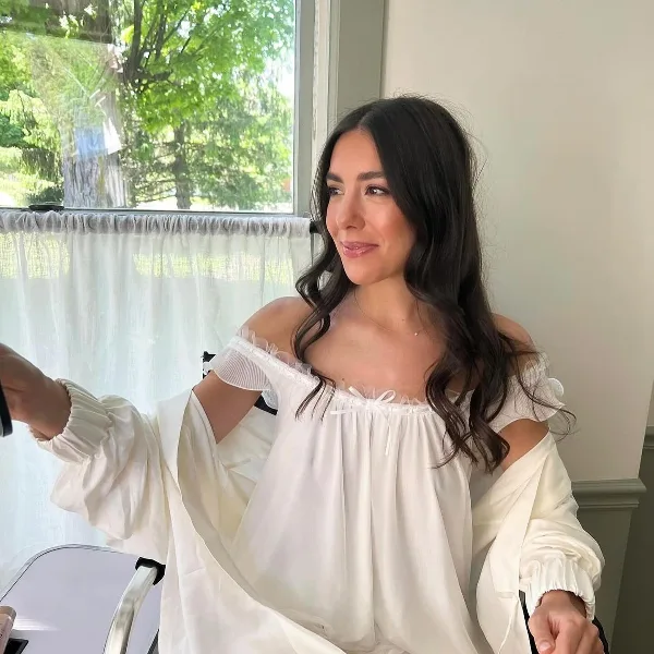 A bride with styled hair and makeup wearing a white robe in a bright room.