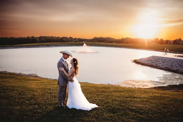 A bride and groom standing on a grassy lawn next to a lake with a fountain at sunset.