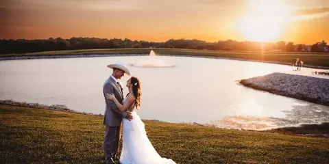 A bride and groom standing on a grassy lawn next to a lake with a fountain at sunset.