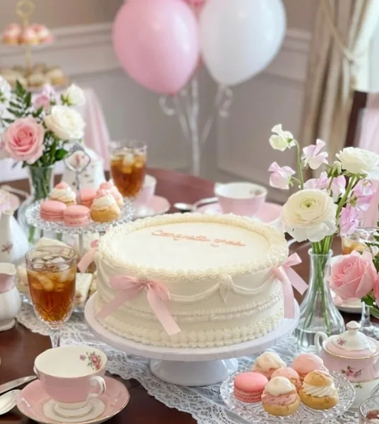 A white frosted cake with pink ribbon accents sits on a pedestal stand, surrounded by macarons, pastries, and floral arrangements on a decorated table.