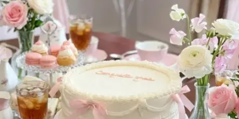 A white frosted cake with pink ribbon accents sits on a pedestal stand, surrounded by macarons, pastries, and floral arrangements on a decorated table.