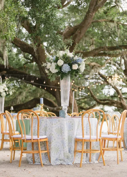 Outdoor wedding reception table with white and blue floral arrangement, wooden chairs, and string lights in the background.
