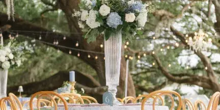 Outdoor wedding reception table with white and blue floral arrangement, wooden chairs, and string lights in the background.