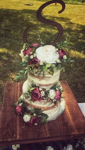 A three-tier semi-naked wedding cake decorated with red and white flowers and a large letter 'S' topper, sitting on a wooden table outdoors.