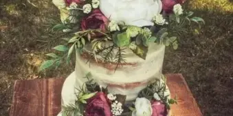 A three-tier semi-naked wedding cake decorated with red and white flowers and a large letter 'S' topper, sitting on a wooden table outdoors.