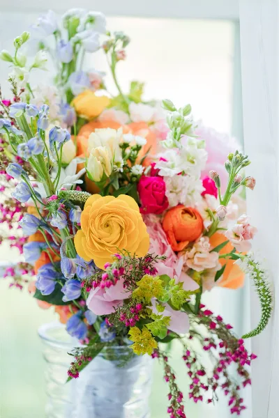 A vibrant floral arrangement featuring yellow ranunculus, pink roses, and blue delphiniums in a clear glass vase.