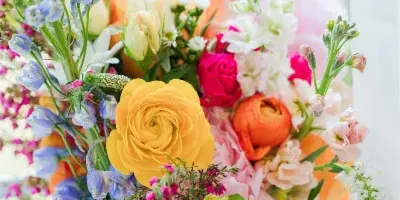 A vibrant floral arrangement featuring yellow ranunculus, pink roses, and blue delphiniums in a clear glass vase.