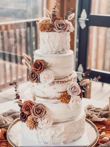 A four-tier white wedding cake decorated with brown and cream roses and dried botanicals, displayed on a gold cake stand.