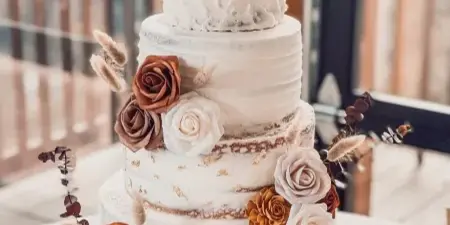 A four-tier white wedding cake decorated with brown and cream roses and dried botanicals, displayed on a gold cake stand.