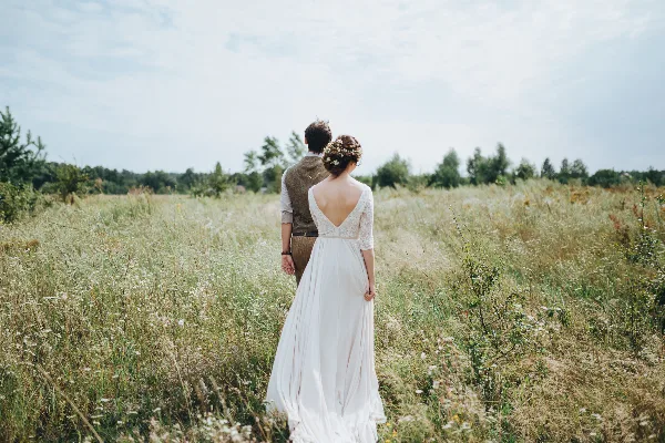 A bride and groom standing in a grassy field with wildflowers, viewed from behind.
