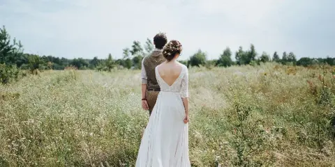 A bride and groom standing in a grassy field with wildflowers, viewed from behind.