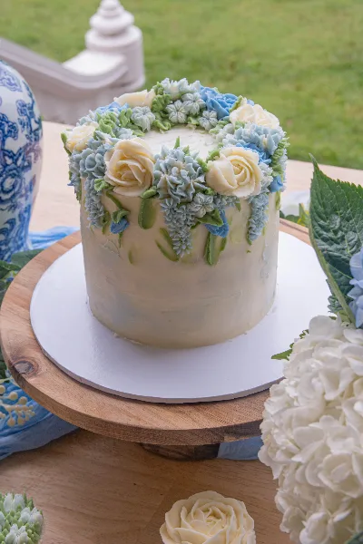 A white frosted cake decorated with blue and cream buttercream flowers on a wooden stand.