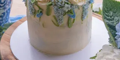 A white frosted cake decorated with blue and cream buttercream flowers on a wooden stand.