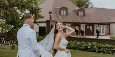 A bride and groom laughing on the lawn in front of a large French provincial style mansion.