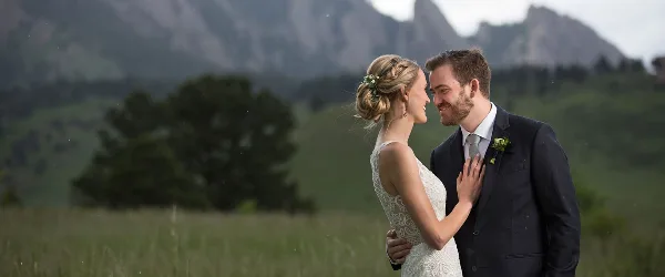 A bride and groom standing in a grassy field with mountains in the background.