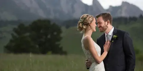 A bride and groom standing in a grassy field with mountains in the background.