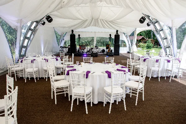 Interior of a clear-frame wedding tent with white table linens, purple napkins, and white chiavari chairs.
