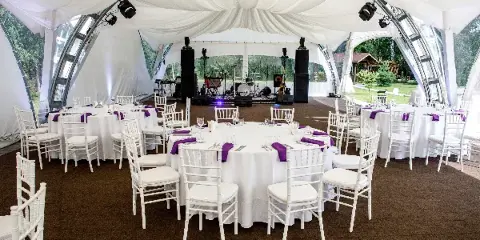 Interior of a clear-frame wedding tent with white table linens, purple napkins, and white chiavari chairs.