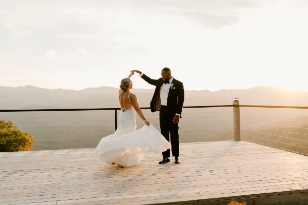 A bride and groom dancing on a wooden deck overlooking a mountain range at sunset.