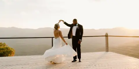 A bride and groom dancing on a wooden deck overlooking a mountain range at sunset.
