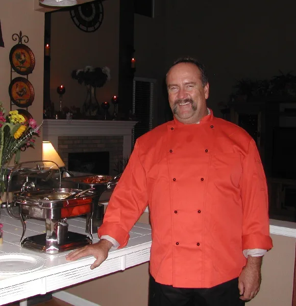 Chef in an orange coat standing next to a buffet table with chafing dishes of food