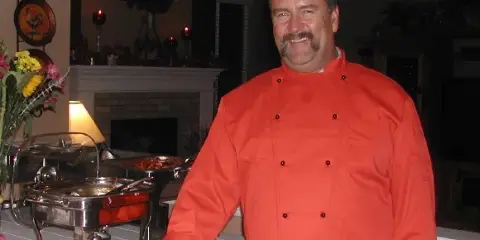 Chef in an orange coat standing next to a buffet table with chafing dishes of food