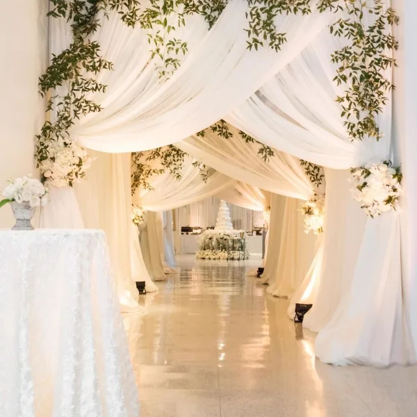 A wedding venue aisle decorated with white draping, greenery, and floral arrangements leading to a wedding cake.